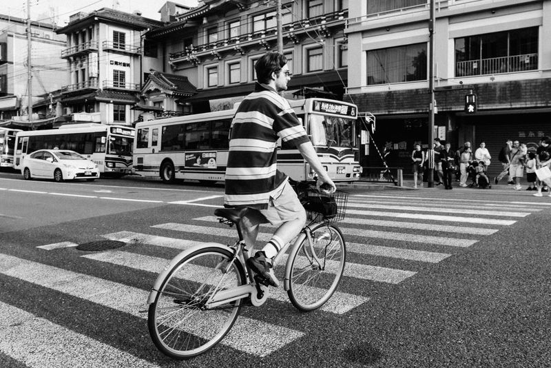 Uomo in bicicletta attraversa un incrocio davanti agli autobus urbani, in bianco e nero, Kyoto