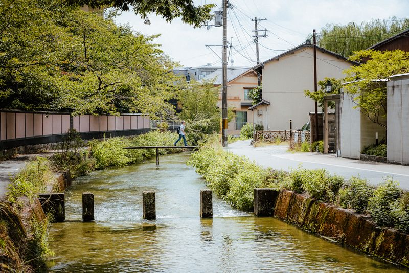 Uomo attraversa una sottile passerella su un canale d'acqua limpida tra le case, Kyoto
