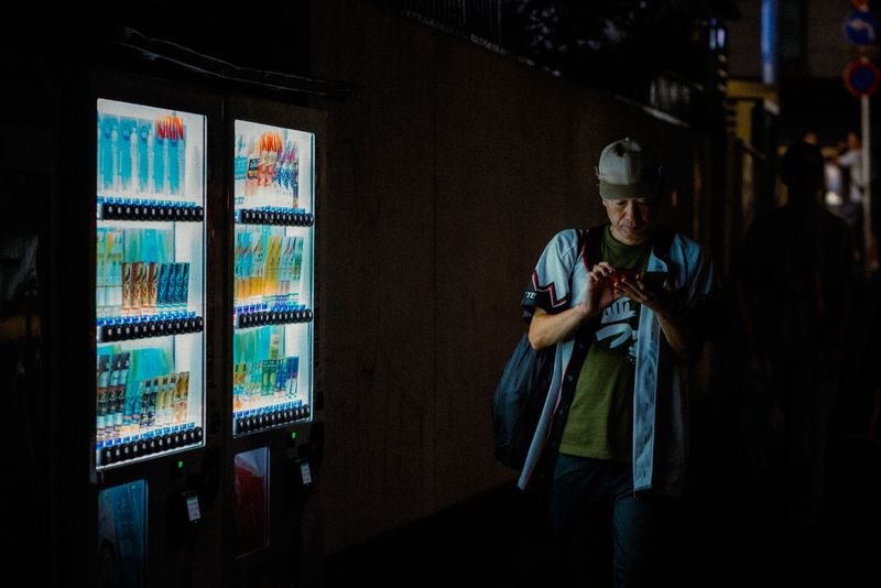 Uomo con cappellino illuminato dal bagliore di due distributori automatici mentre guarda il telefono, di notte, Kyoto