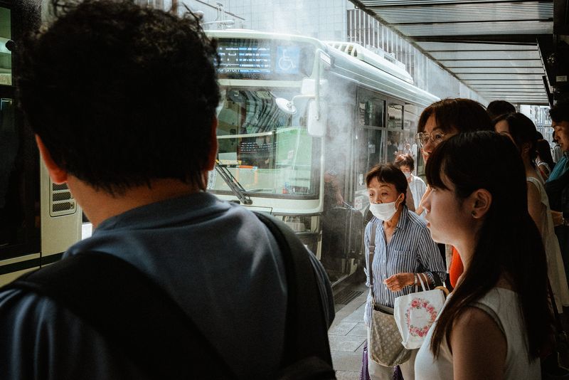 Passeggeri in attesa alla fermata dell'autobus sotto il getto di un nebulizzatore estivo, Kyoto