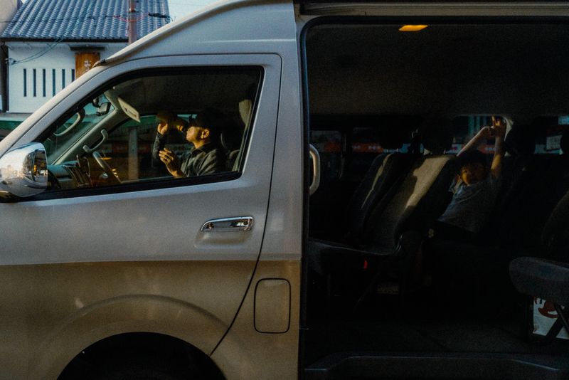 Autista beve al volante di un van mentre un bambino gioca sul sedile posteriore, luce del tramonto, Kyoto
