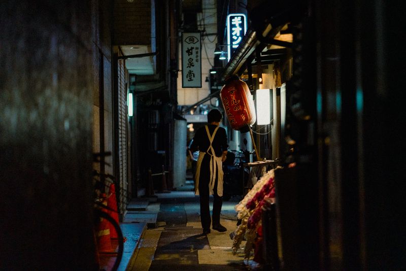 Un lavoratore in grembiule in un vicolo buio di Gion accanto a una lanterna rossa, Kyoto