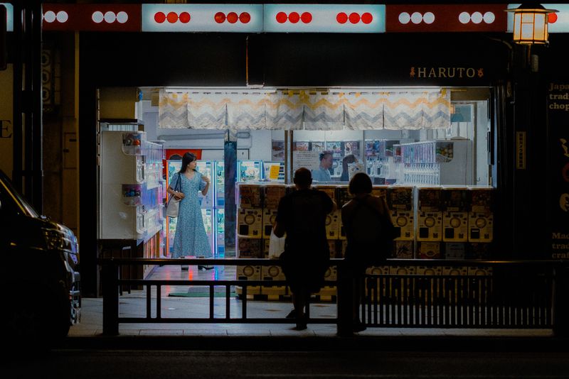 Donna in abito azzurro tra le macchine di capsule gachapon del negozio Haruto, osservata da due passanti, di notte, Kyoto