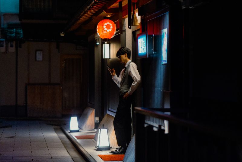 Giovane in gilet controlla il telefono sotto una lanterna rossa in un vicolo di Gion, di notte, Kyoto
