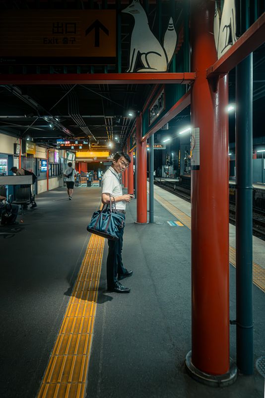 Un uomo con la borsa si volta sulla banchina della stazione di Fushimi Inari sotto l'insegna con la volpe, di notte, Kyoto