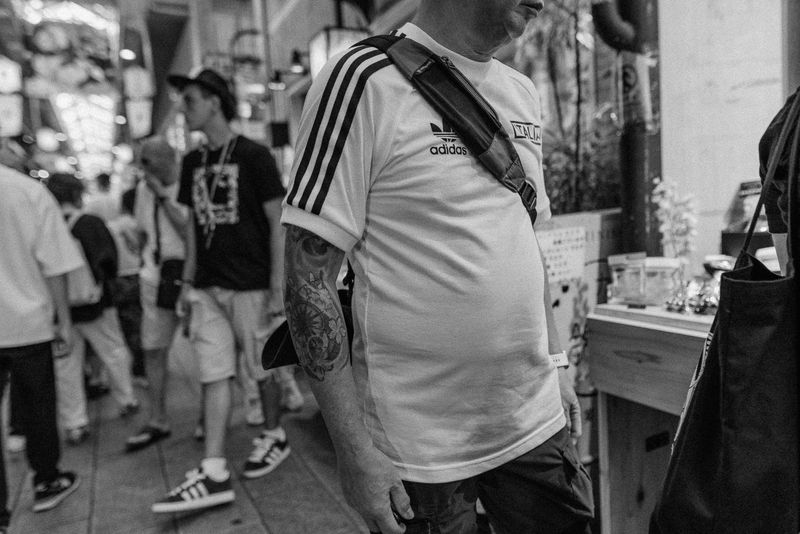 Uomo con maglia Adidas Italia e braccio tatuato tra la folla di un mercato, in bianco e nero, Kyoto