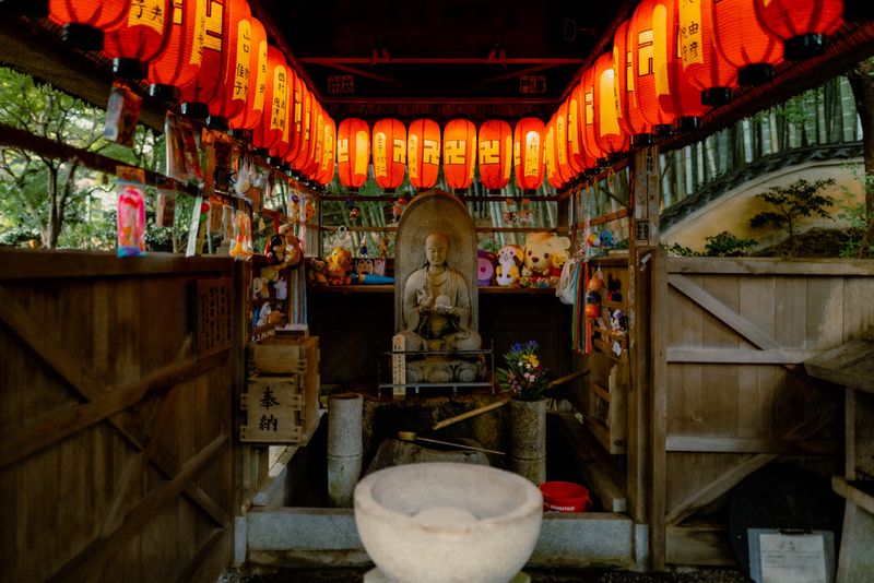 Piccola cappella con statua di pietra tra lanterne rosse e peluche votivi, dintorni di Kyoto