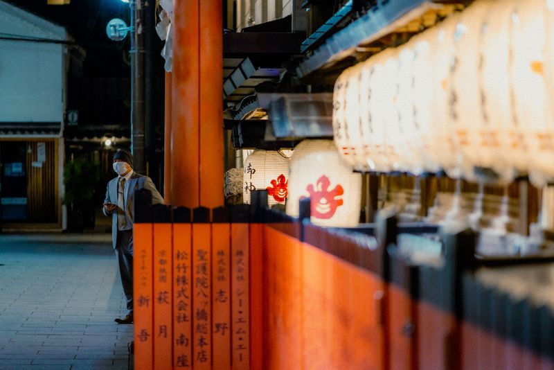 Uomo con mascherina appoggiato a un palo vermiglio accanto a una fila di lanterne bianche accese, di notte, Kyoto