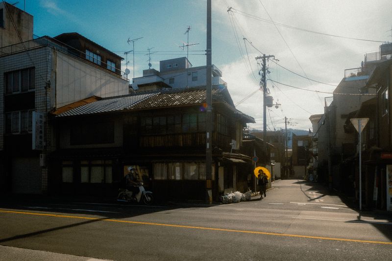 Persona con ombrello giallo cammina in una via silenziosa tra le vecchie case, uno scooter all'angolo, Kyoto