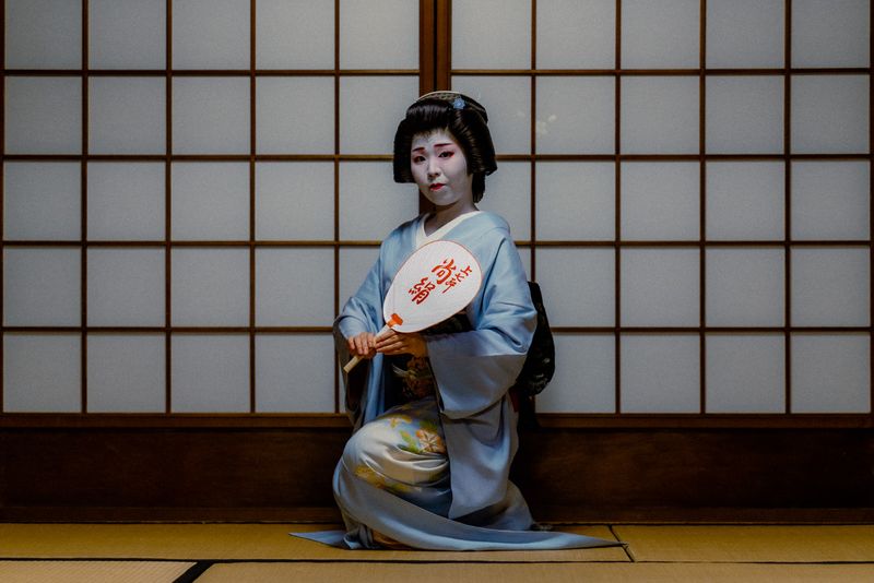 Una maiko inginocchiata sul tatami con un ventaglio uchiwa davanti a un fusuma di carta, Kyoto