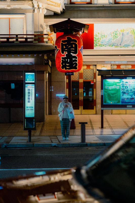 La grande lanterna rossa del teatro Minamiza sopra una donna alla fermata del bus, di notte, Kyoto