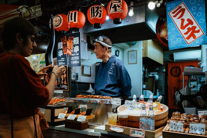 Venditore in blu al banco degli spiedini di anguilla mentre una cliente fotografa il menù, mercato Nishiki, Kyoto