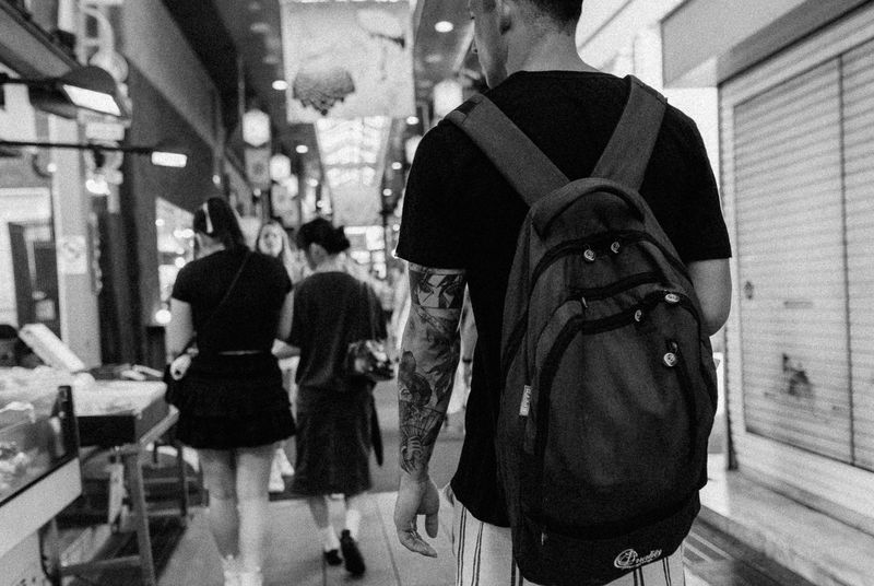 Uomo di spalle con zaino e braccio tatuato lungo la galleria del mercato Nishiki, in bianco e nero, Kyoto