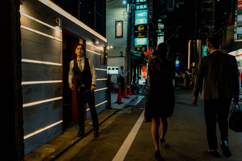 Un uomo in gilet attende sull'uscio mentre una coppia passa in un vicolo al neon, di notte, Kyoto