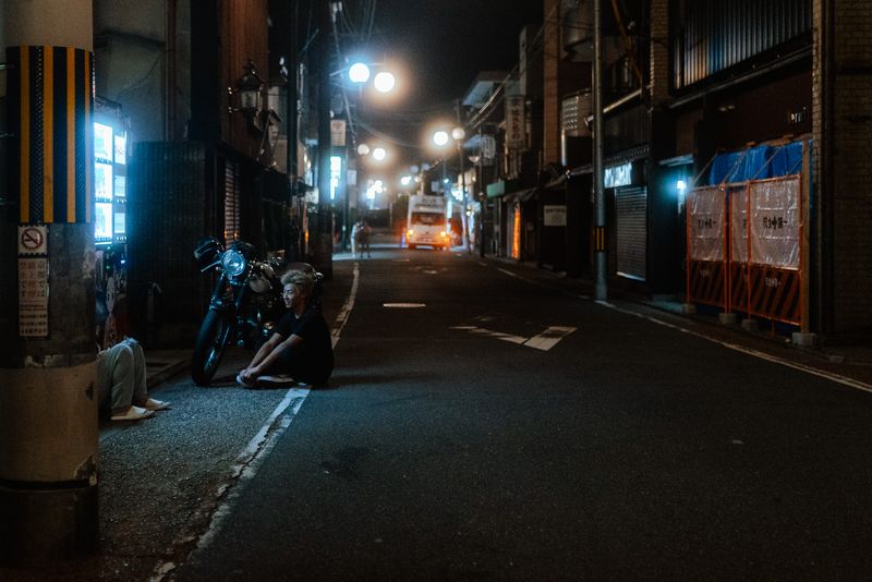 Un giovane seduto a terra accanto a una moto café racer in una via deserta, di notte, Kyoto