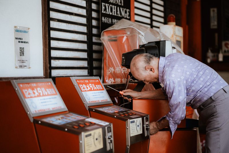Anziano ripara una macchina di medaglie commemorative accanto a un cambio valuta, Okazaki, Kyoto