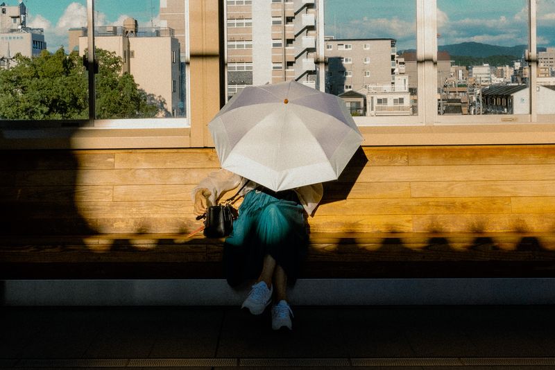 Donna con parasole seduta su una panca di legno davanti a una grande vetrata sulla città, Kyoto