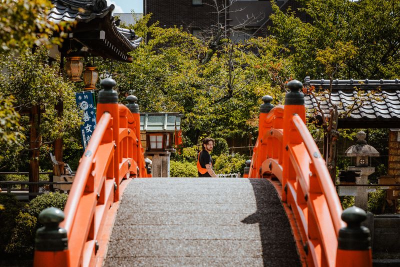 Un uomo con gilet arancione oltre un ponte di legno rosso in un giardino di santuario, Kyoto