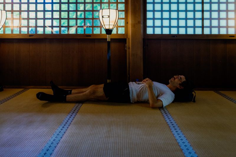 Un uomo disteso sul tatami riposa sotto una lanterna, in una sala del tempio, Kyoto