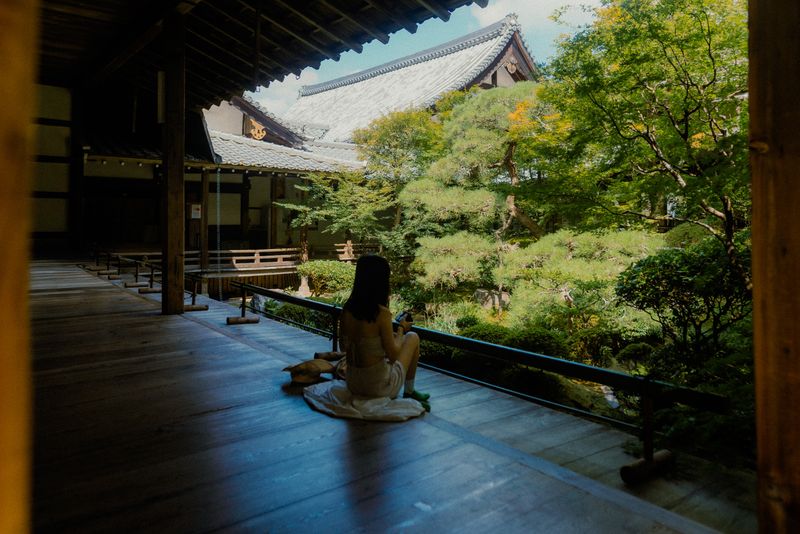 Donna seduta sulla veranda di un tempio osserva il giardino con i tetti e i pini, Kyoto
