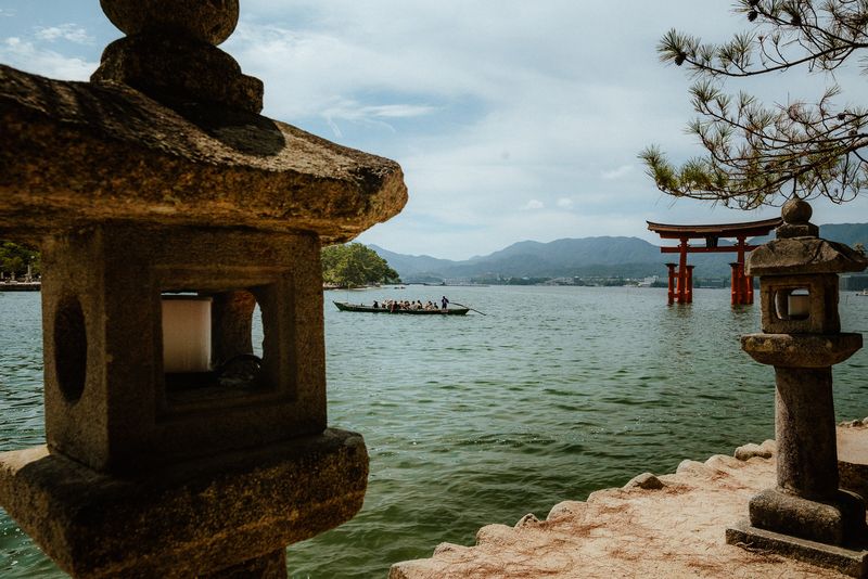 Lanterne di pietra in primo piano e una barca di turisti diretta verso il torii galleggiante, Miyajima