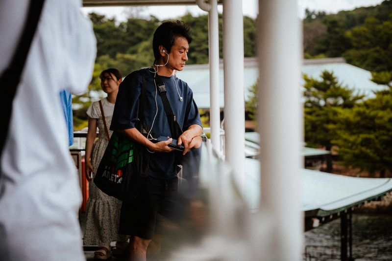 Passeggeri sul ponte del traghetto per Miyajima, un uomo con gli auricolari guarda verso l'isola