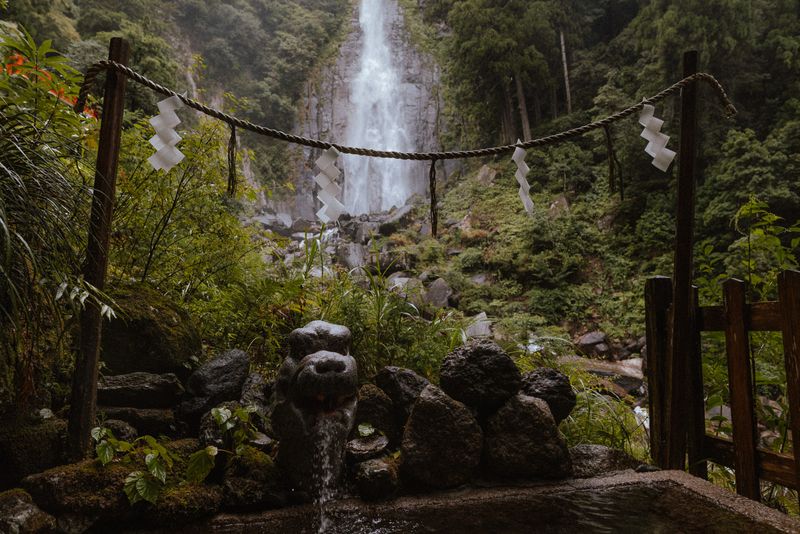 Fontana purificatrice a testa di drago sotto la corda shimenawa, con la cascata di Nachi sullo sfondo