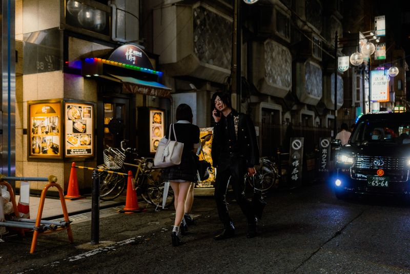 Un uomo al telefono e una donna si incrociano sul marciapiede mentre un taxi svolta nel vicolo, di notte, Osaka
