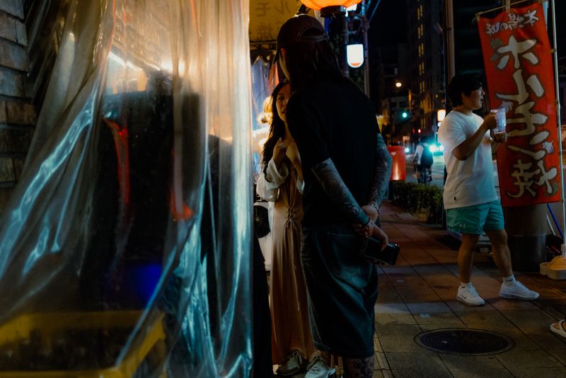 Coppia appoggiata a una tenda di plastica illuminata mentre un ragazzo beve accanto al banner rosso, di notte, Osaka