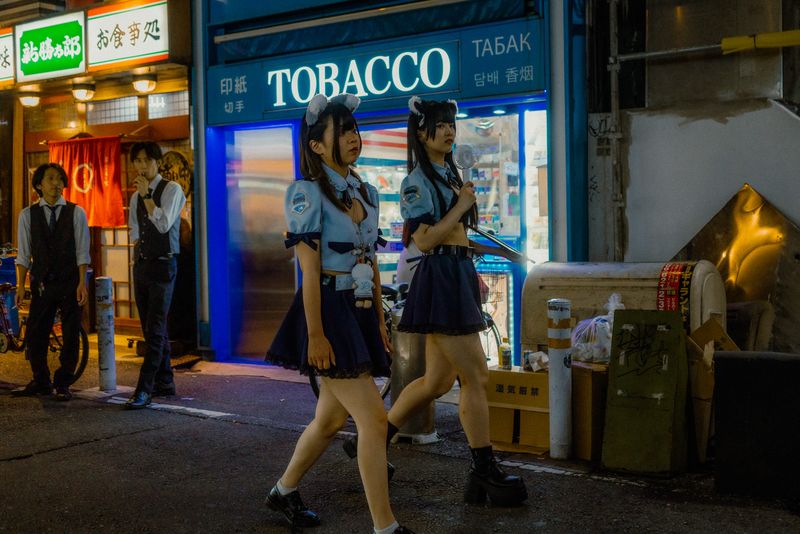 Due ragazze in costume passano davanti a una tabaccheria mentre due camerieri in gilet guardano dalla soglia, Osaka