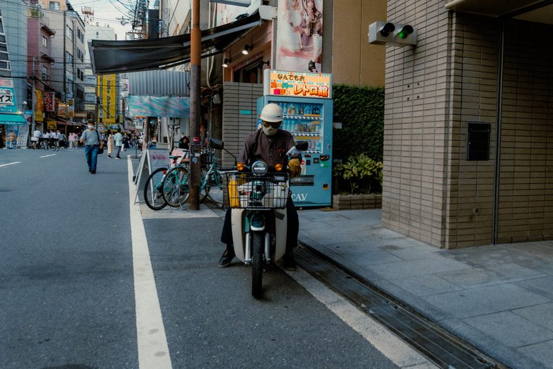 Uomo con casco e mascherina in sella a un Honda Super Cub accanto a un distributore, Osaka