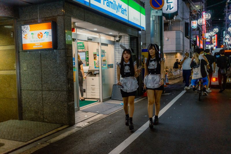 Due ragazze in costume da cameriera camminano lungo la strada davanti a un FamilyMart, di notte, Osaka