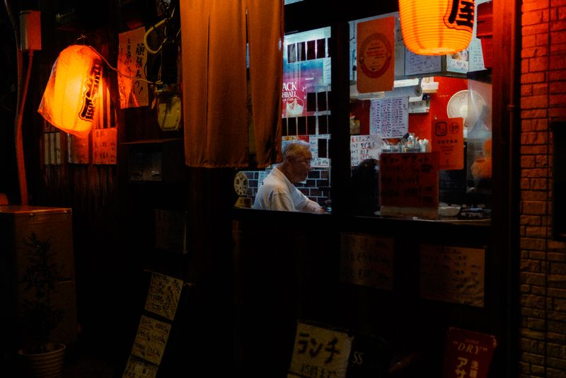 Anziano seduto al bancone di un izakaya visto dalla finestra tra due lanterne rosse, Osaka