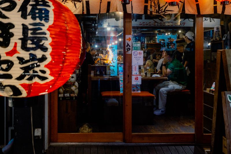 Fiammata alla griglia dentro un izakaya visto oltre una grande lanterna rossa in primo piano, Osaka