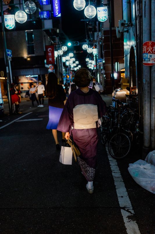 Donna in kimono cammina di spalle sotto la fila di lampioni tondi di una via di Shinsaibashi, di notte, Osaka