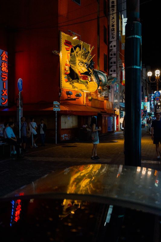 Il drago del Kinryū Ramen all'angolo, una ragazza sola sul marciapiede e i neon riflessi sul cofano di un'auto, Osaka