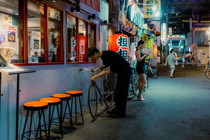 Uomo che chiude la bicicletta accanto a una fila di sgabelli arancioni, vicolo di Kyōbashi di notte, Osaka