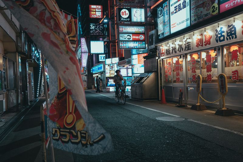 Ciclista che attraversa la strada tra le insegne al neon dei locali di Kyōbashi, di notte, Osaka