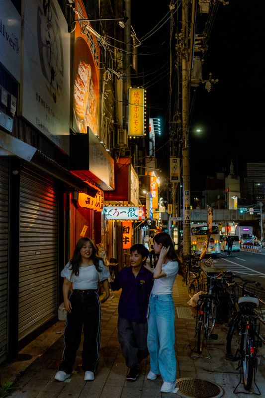 Tre ragazzi camminano ridendo sul marciapiede tra le insegne di yakiniku, Kyōbashi di notte, Osaka