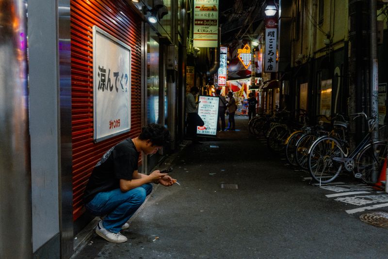 Uomo accovacciato con sigaretta e telefono in un vicolo di izakaya illuminato, Kyōbashi, Osaka