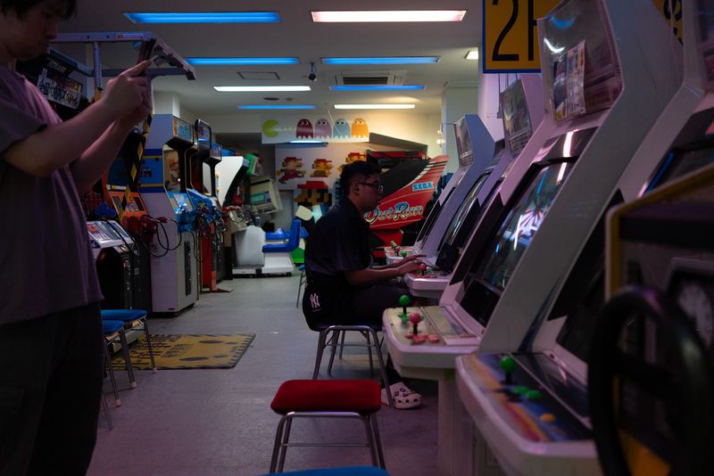 Ragazzo seduto a un cabinato mentre un altro fotografa, sala giochi con Out Run e Pac-Man alle pareti, Osaka