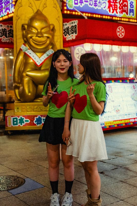 Due ragazze in maglietta verde posano davanti alla statua dorata di Billiken e alle insegne al neon di Shinsekai, Osaka