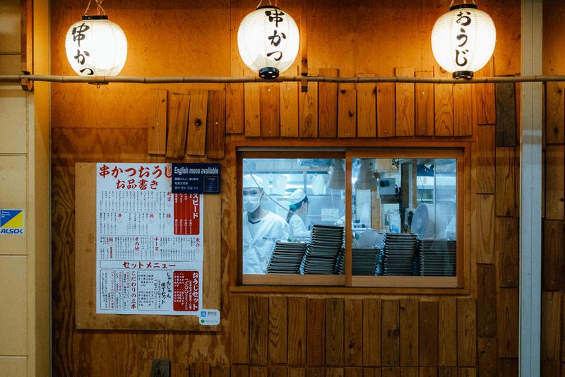 Facciata in legno di un locale di kushikatsu con tre lanterne accese e i cuochi visti dalla finestra, Shinsekai, Osaka