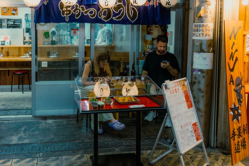 Coppia di turisti al tavolo esterno di un locale di kushikatsu accanto all'insegna Ninja, Shinsekai, Osaka