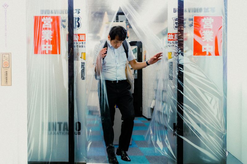 Uomo in camicia che esce da una tenda di plastica trasparente sotto le insegne 営業中, Osaka