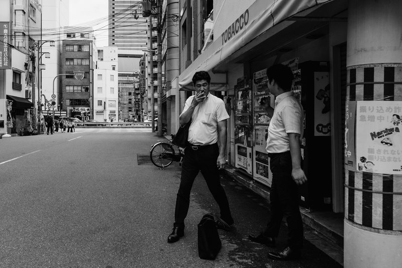Uomo in camicia fuma davanti a un negozio di tabacchi, in bianco e nero, Osaka