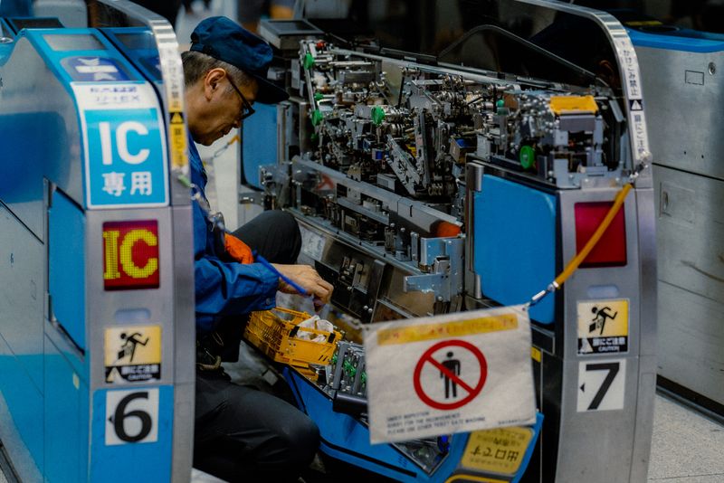 Tecnico in divisa blu al lavoro dentro un tornello aperto della stazione, ingranaggi a vista, Osaka