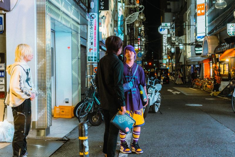 Tre ragazzi conversano all'angolo di una via commerciale illuminata, di notte, Osaka