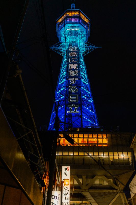 La torre Tsūtenkaku illuminata di blu vista dal basso tra i cavi e le insegne di Shinsekai, Osaka
