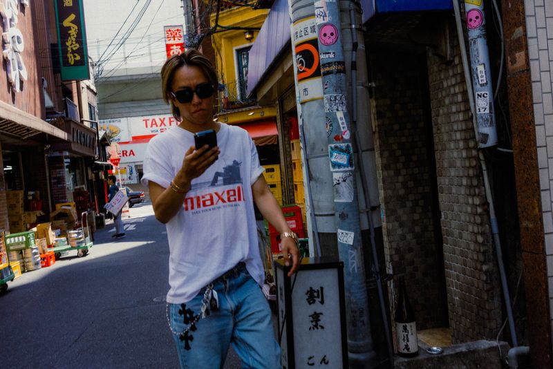 Ragazzo biondo con occhiali da sole e maglietta Maxell guarda il telefono in un vicolo di consegne, Osaka